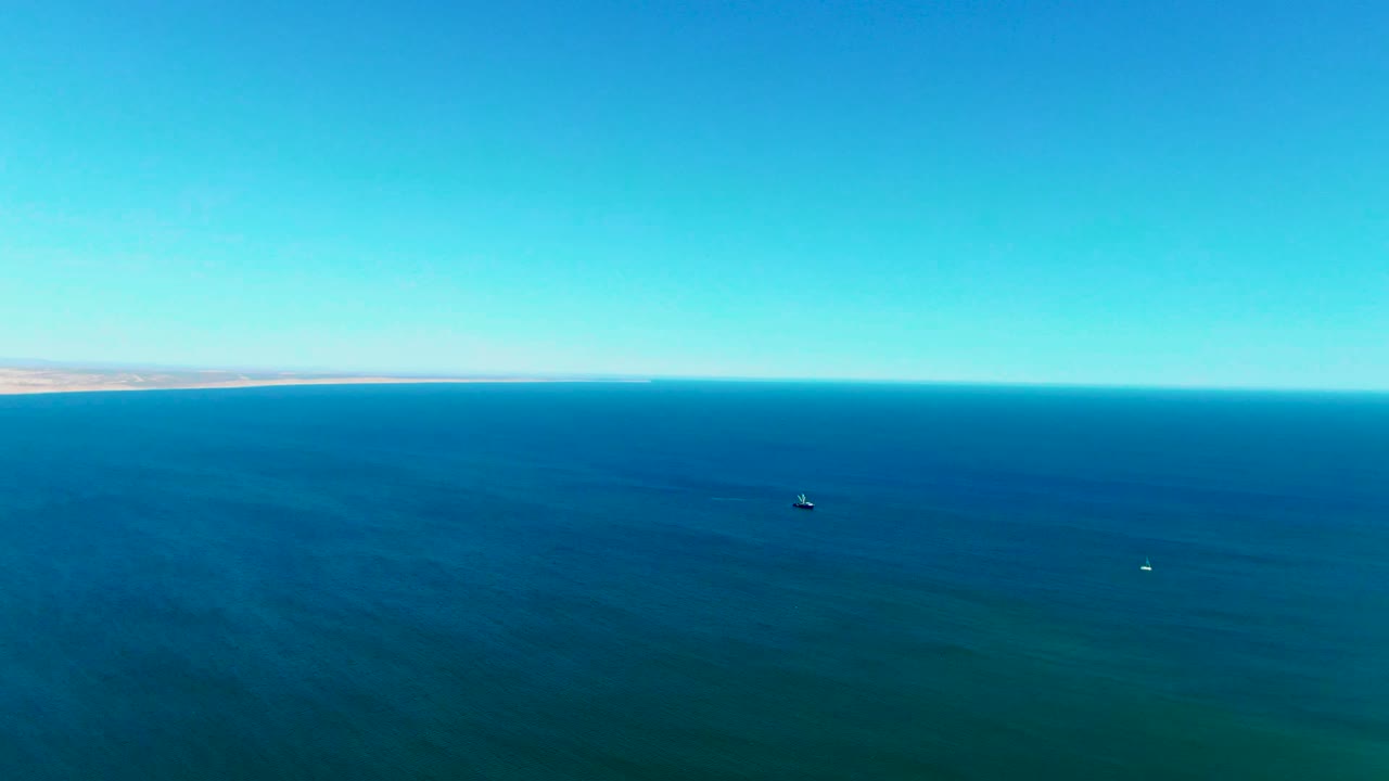 Wide aerial view over blue ocean near San Juanico, Baja California Sur, with a faint shoreline on the horizon, small boats on the water, and no visible people.