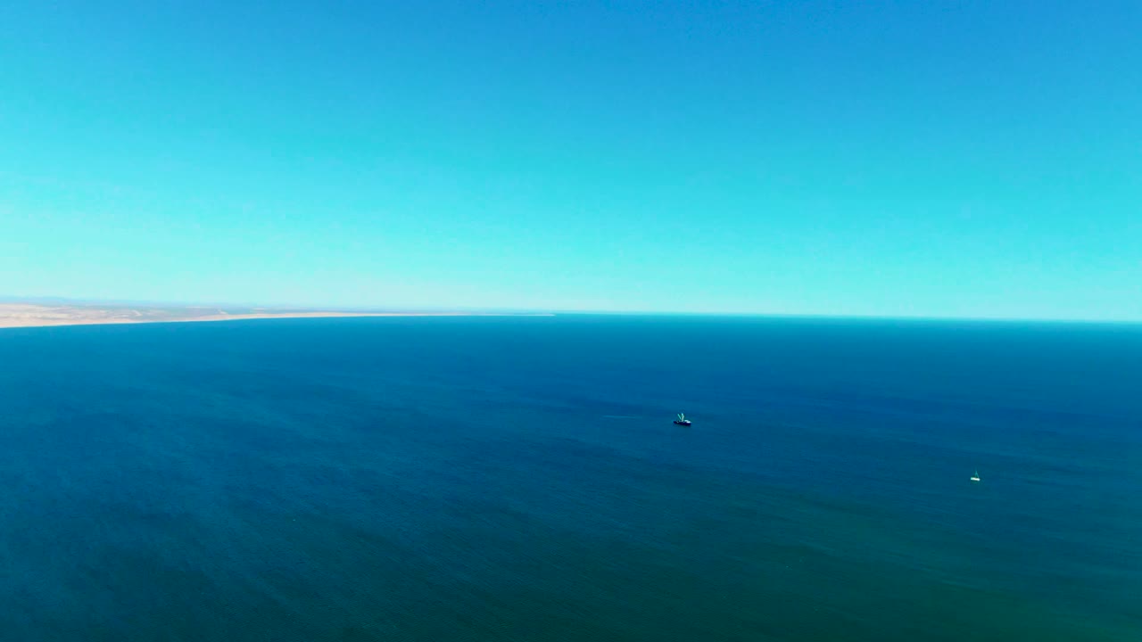 Wide drone-style view of San Juanico ocean water with a straight horizon, pale distant land at left, small vessels on the sea, and no visible people.