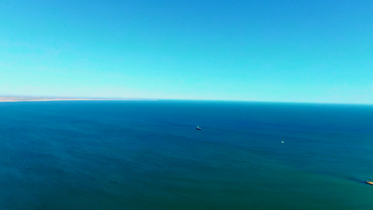 Wide aerial view over the blue ocean near San Juanico, Baja California Sur, with a thin shore on the horizon, small boats on the water, and no visible people.