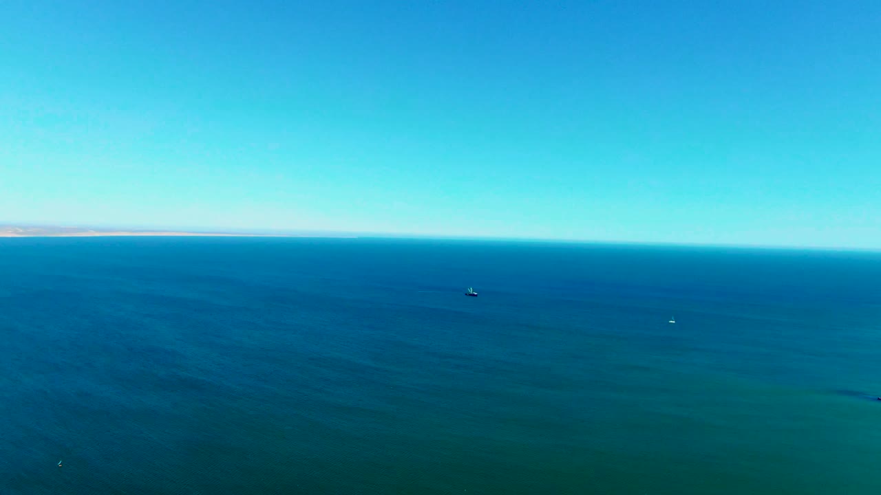 Aerial view over blue-green ocean near San Juanico, Baja California Sur, with a clear sky, thin distant land on the horizon, small floating objects on the water, and no visible people.