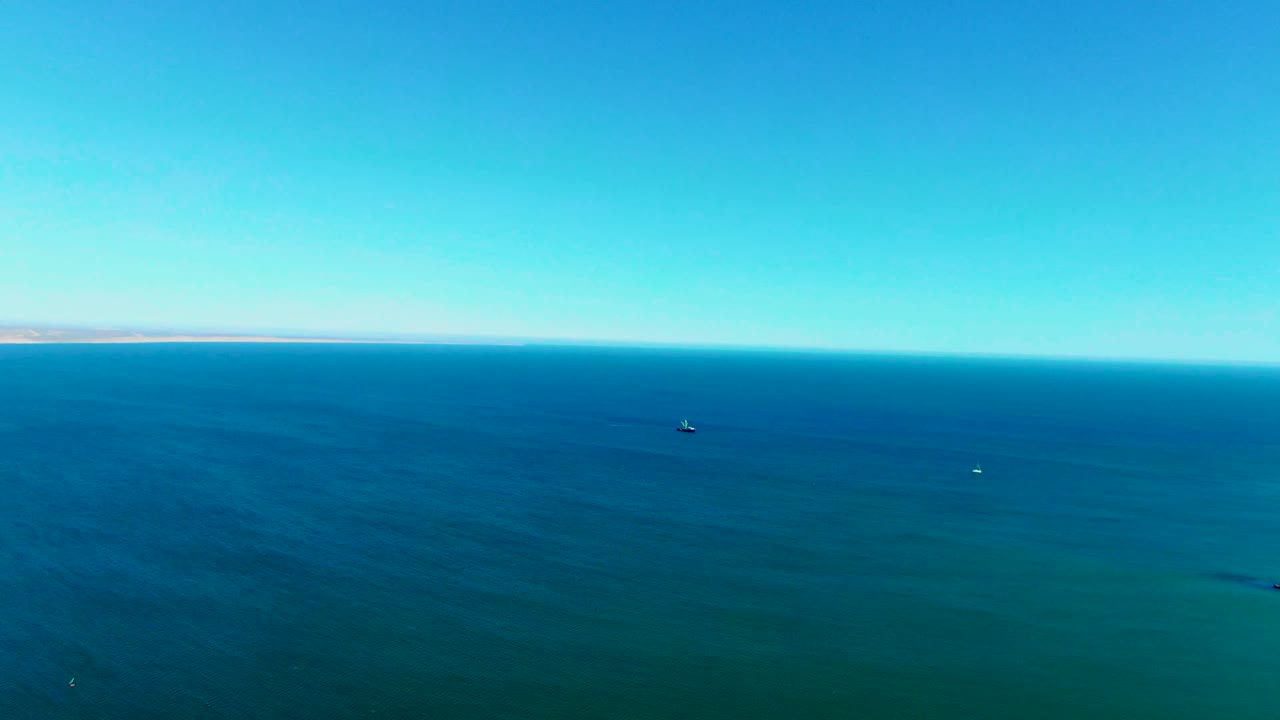 Wide aerial frame over San Juanico ocean water, showing small buoy-like points on the surface, a faint land strip along the horizon, clear blue sky, and no visible people.