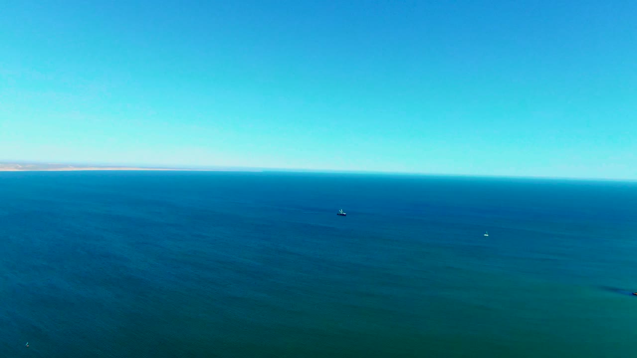 Aerial ocean view in Baja California Sur with blue water filling most of the frame, a narrow distant shoreline at the horizon, scattered small floating markers, and no visible people.