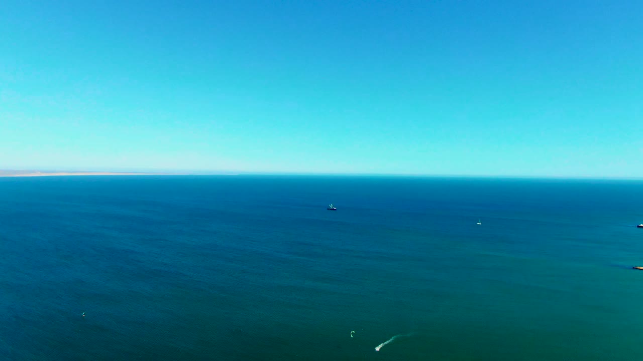Wide aerial frame over blue-green ocean near San Juanico, Baja California Sur, with small distant boats, a clear horizon, faint land, and no visible people.