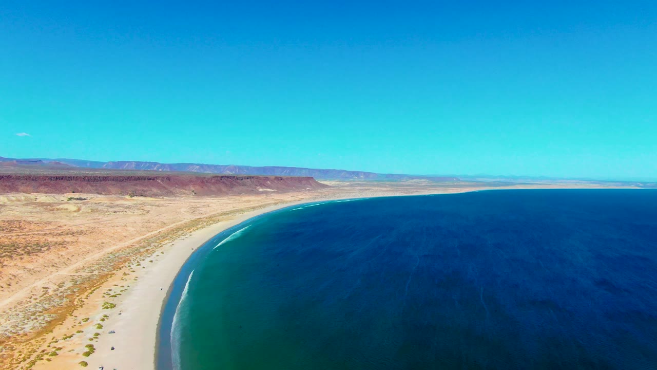 Aerial view over San Juanico, Baja California Sur, showing a broad sandy beach, deep blue water, arid terrain, distant low mountains, and no visible people.