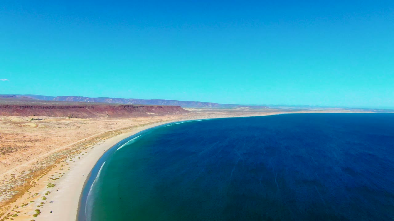 Aerial coastline with beach, green shallows, blue water, and desert plateau.