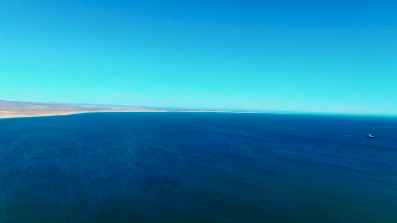 Wide aerial view near San Juanico, Baja California Sur, looking across open blue ocean with a distant desert shoreline, a small boat to the right, and no visible people.