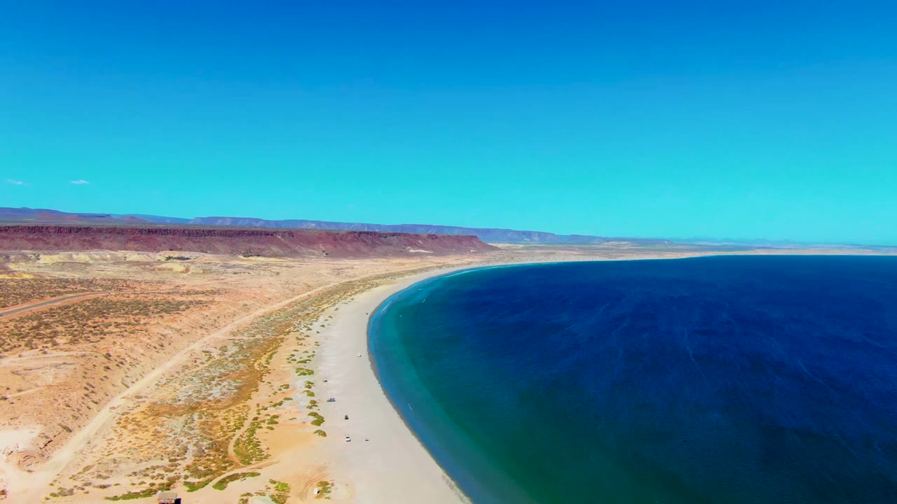 Aerial frame over San Juanico, Baja California Sur, with sandy beach and desert scrub on the left, deep blue ocean on the right, distant mesa terrain, and no visible people.