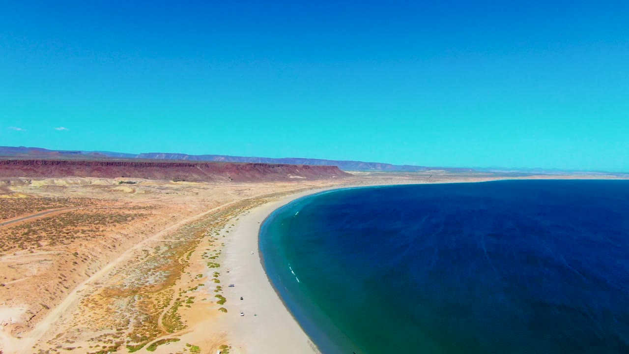 Aerial view in San Juanico showing the beach arc slightly farther along the coast, with turquoise water near shore, open desert flats, distant reddish hills, and no visible people.