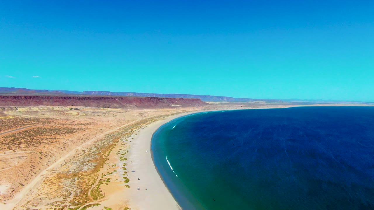 Aerial San Juanico coastline frame with a small white wave beginning to show near the beach, dark blue open water, pale sand, desert vegetation, and no visible people.