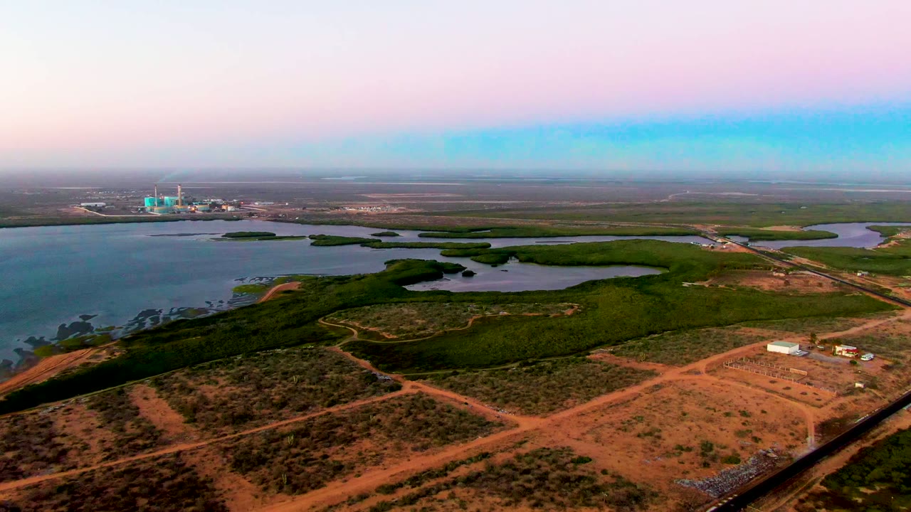 High aerial view over San Carlos, Baja California Sur, with reddish dirt roads in the foreground, green vegetation around shallow water, distant industrial structures on the horizon, and no visible people.
