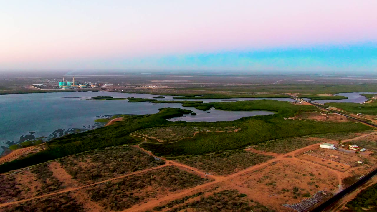Aerial landscape with intersecting dirt tracks, green shoreline vegetation, water channels, and distant buildings.