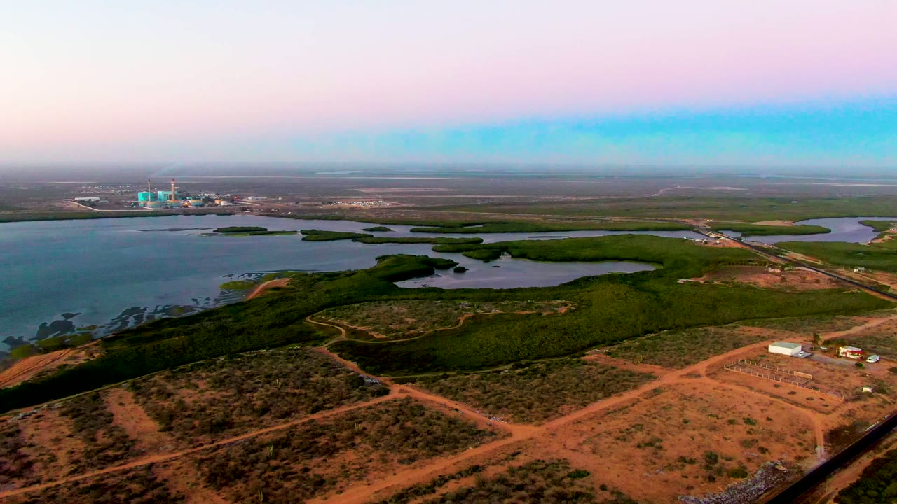 Aerial frame over San Carlos, Baja California Sur, with a small white structure at lower right, reddish dirt roads, scrub-covered ground, water and green vegetation beyond, and no visible people.