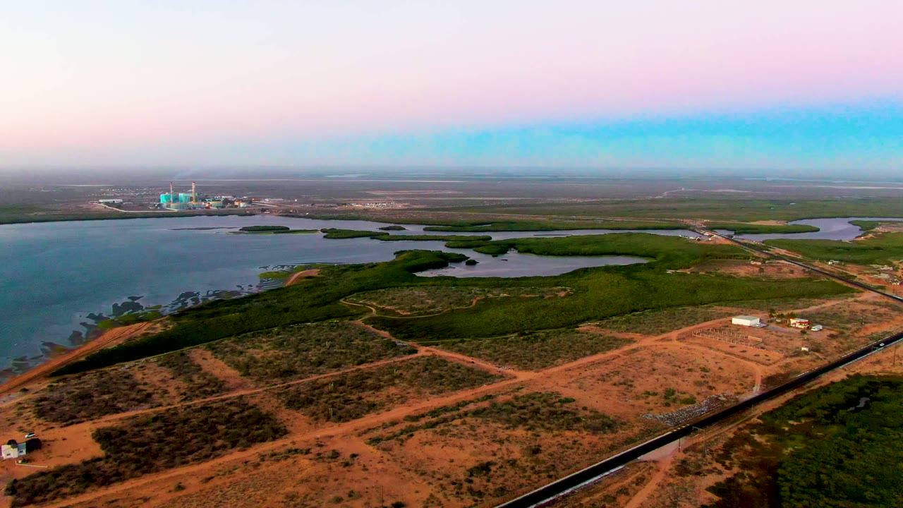 Aerial frame from San Carlos, Baja California Sur showing reddish dirt tracks, green wetland areas, open water, distant structures, and no visible people at the start of the clip.