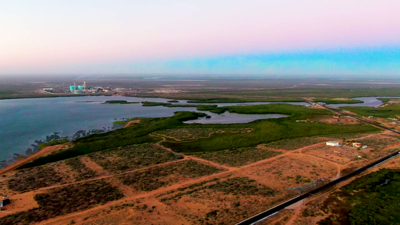 Wide drone view over reddish ground, wetland channels, and distant structures near the horizon.