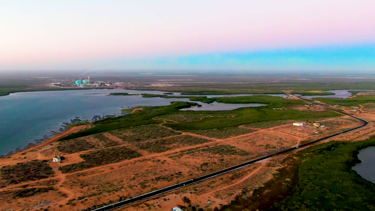 Aerial frame from San Carlos, Baja California Sur showing a railway along coastal water, green wetlands, reddish dirt roads, distant buildings, and no visible people.