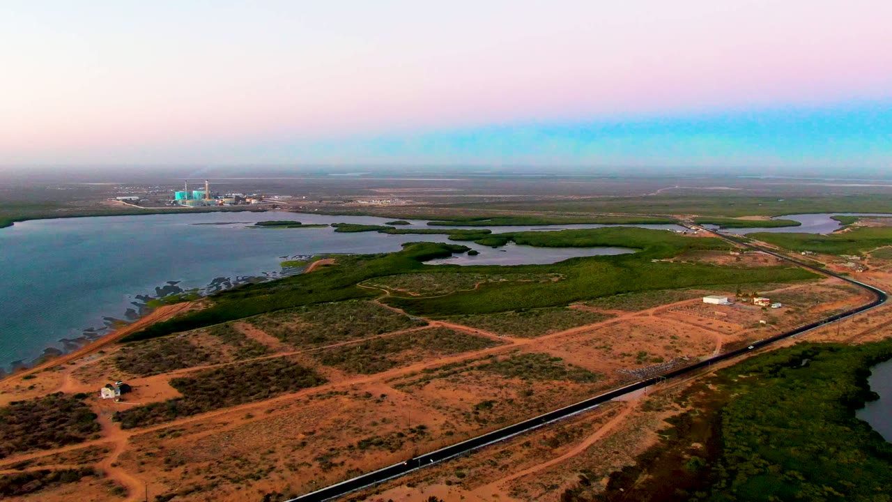 Aerial frame over San Carlos, Baja California Sur with reddish terrain in the foreground, the coastal railway near the bottom edge, wetlands and water beyond, and no visible people.