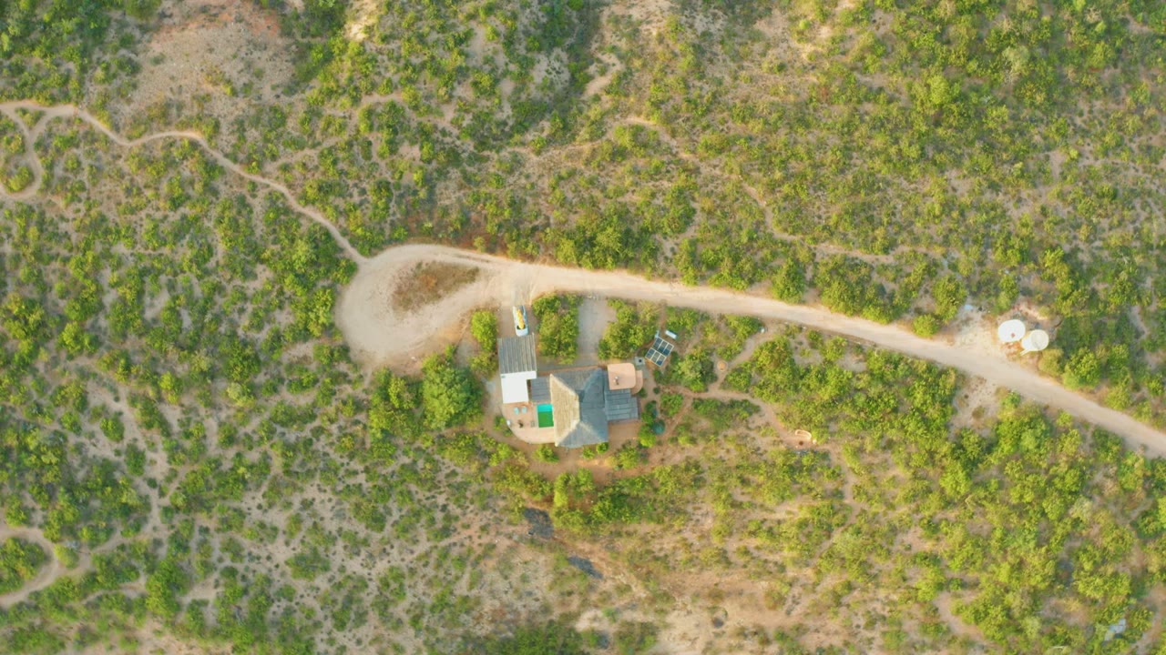 Overhead frame from Cabo Pulmo, Baja California Sur showing a pale dirt road curving past roofed structures and green-brown hillside scrub, with no visible people.
