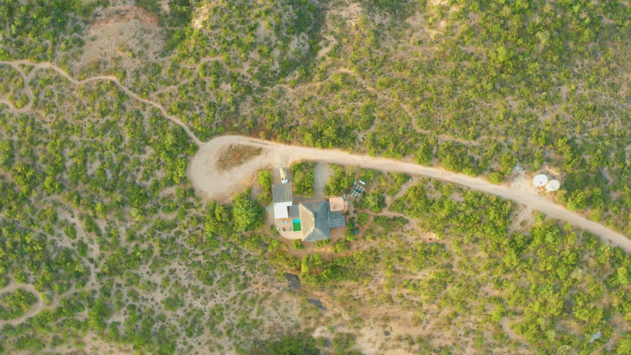 At 0:02, the drone view over Cabo Pulmo shows the road stretching across the top of frame while the buildings and small car sit below among uneven green vegetation, with no visible people.