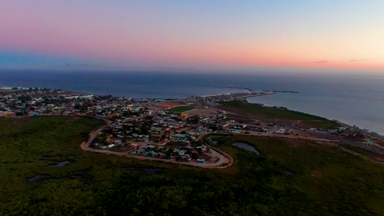 Wide aerial view of San Carlos, Baja California Sur at dusk, with dark green foreground, a small coastal town, ocean horizon, and no visible people.