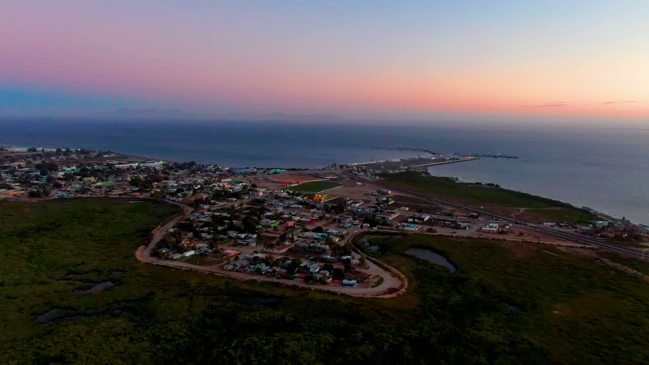 High aerial view of San Carlos, Baja California Sur with town roofs, road corridors, dark green land, a small shoreline area, and no visible people.