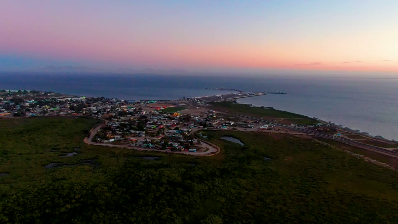 Aerial dusk view over San Carlos, Baja California Sur, with dark green foreground, clustered town buildings, a winding coastal road, ocean water, pink-blue sky, and no visible people.