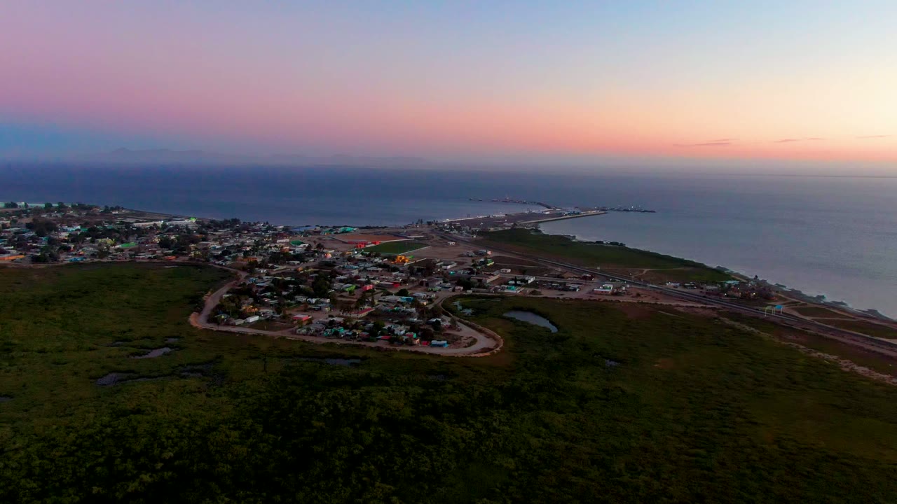 Wide aerial view of town buildings near the ocean under a pink and blue dusk sky.