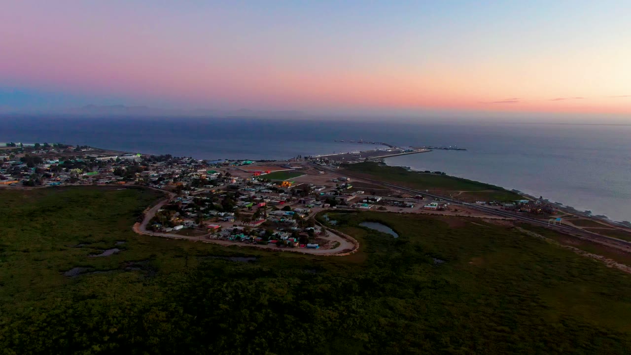 Aerial view of San Carlos at dusk with the settlement spread across the left side, a road and shoreline crossing the middle, ocean in the background, and no visible people.
