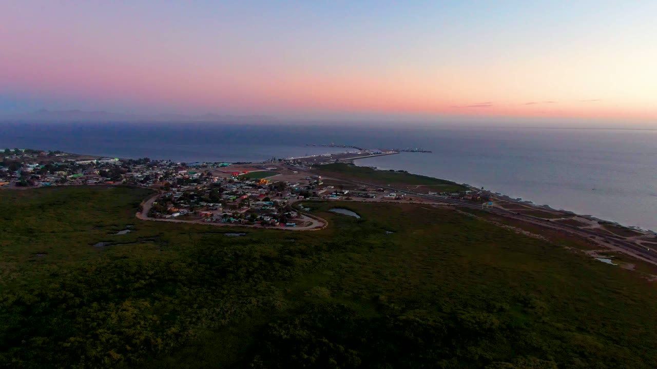 Aerial dusk view over San Carlos, Baja California Sur, with dark green vegetation in front, a coastal town and winding road below, ocean beyond, and no visible people.