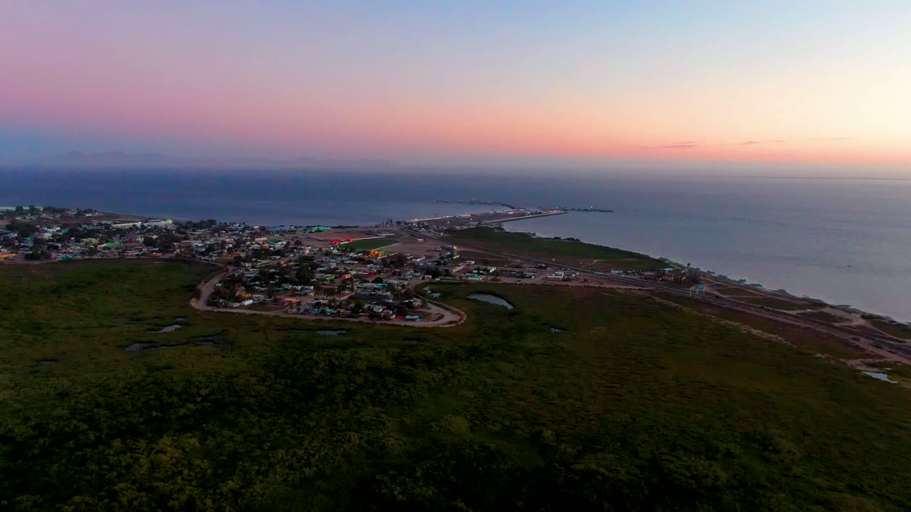 A high aerial frame over San Carlos, Baja California Sur shows town rooftops, small evening lights, a winding road, ocean water, and a pink-purple sky with no visible people.