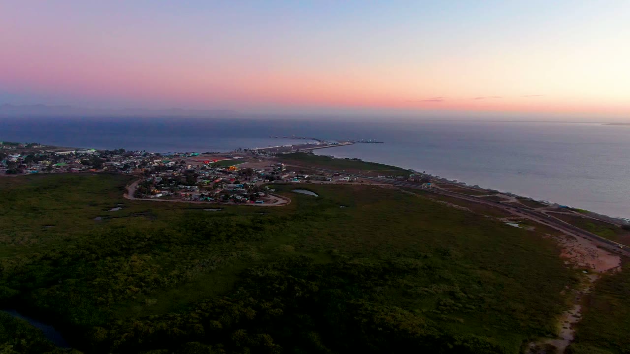 Aerial dusk view over San Carlos, Baja California Sur, with dark green foreground land, a small coastal town at left, ocean at right, pink-blue sky, and no visible people.