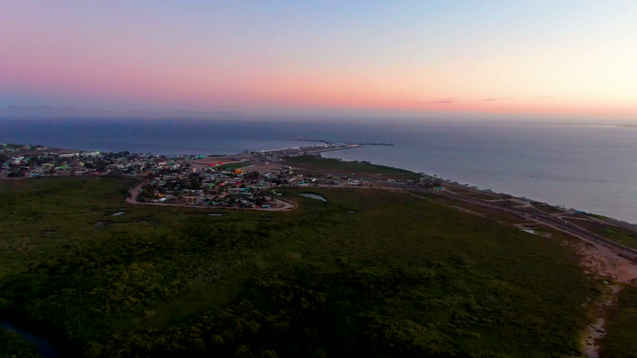 Wide aerial view of coastal buildings, green foreground, and ocean at sunset.
