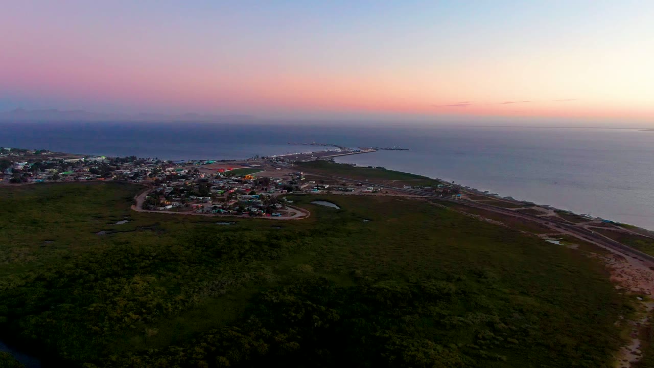 Aerial view over San Carlos, Baja California Sur, with the town slightly left of center, dark vegetation in the foreground, shoreline infrastructure, open ocean, and no visible people.