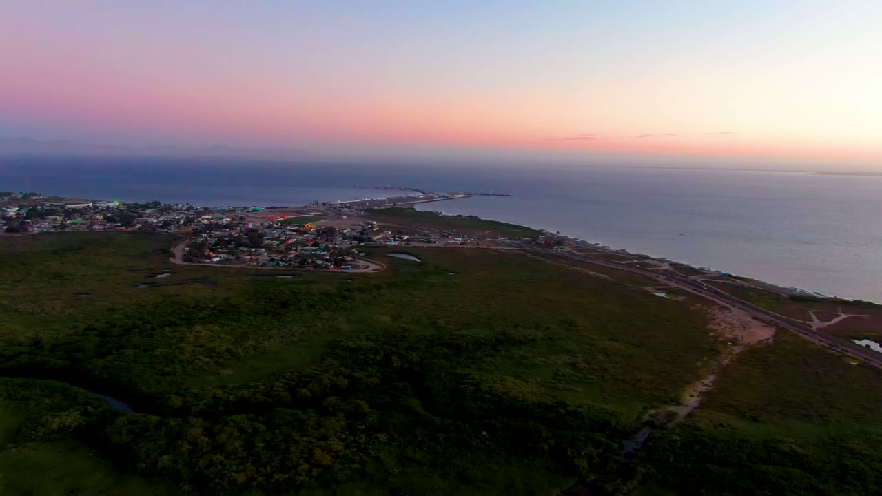 Opening aerial frame over San Carlos, Baja California Sur, with dark green fields in front, a coastal village near the shore, ocean beyond, pink dusk light, and no visible people.