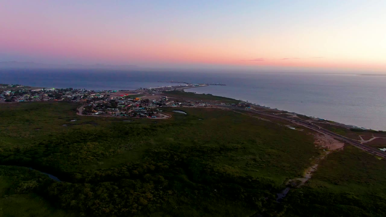 Aerial dusk view over San Carlos, Baja California Sur, with broad green land in the foreground, clustered buildings near the water, pale ocean on the right, and no visible people.