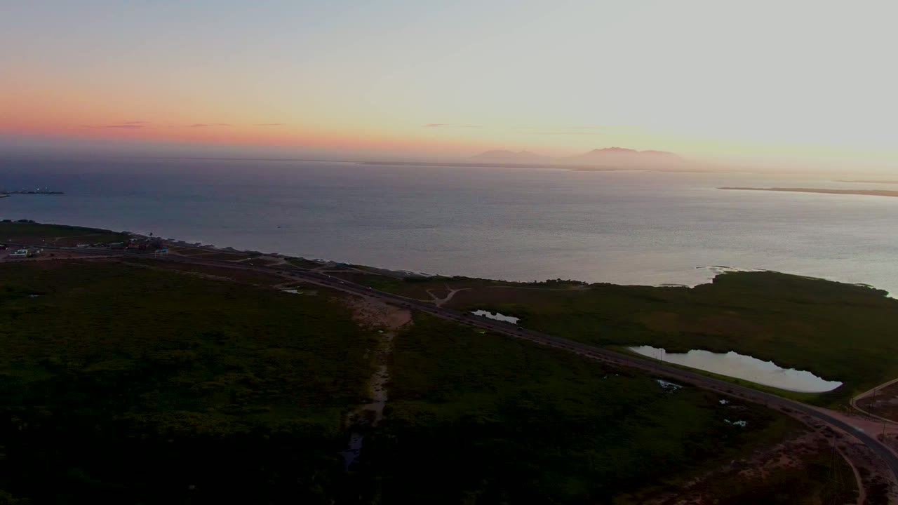 Aerial view in San Carlos, Baja California Sur, with a road crossing dark foreground land toward the ocean under a pastel sunset sky and no visible people.