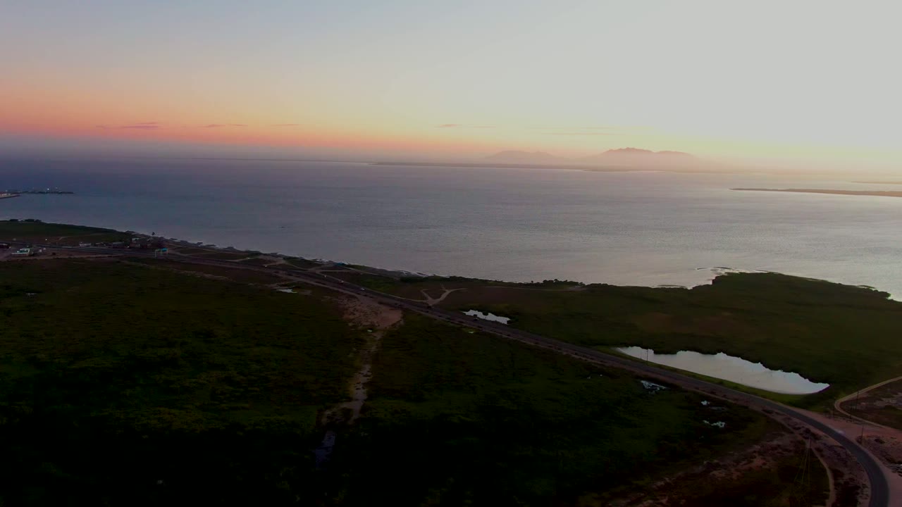 Wide aerial frame of San Carlos, Baja California Sur, with the road cutting across green coastal terrain beneath a soft orange and blue sky, with no visible people.
