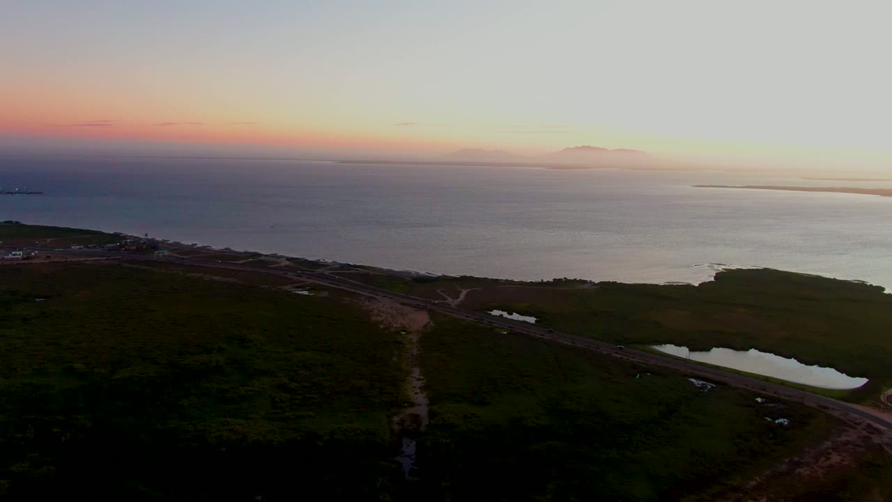 A wide San Carlos, Baja California Sur drone frame shows the road cutting across dark lowland, the small lagoon reflecting sky color, and the ocean horizon with no visible people.