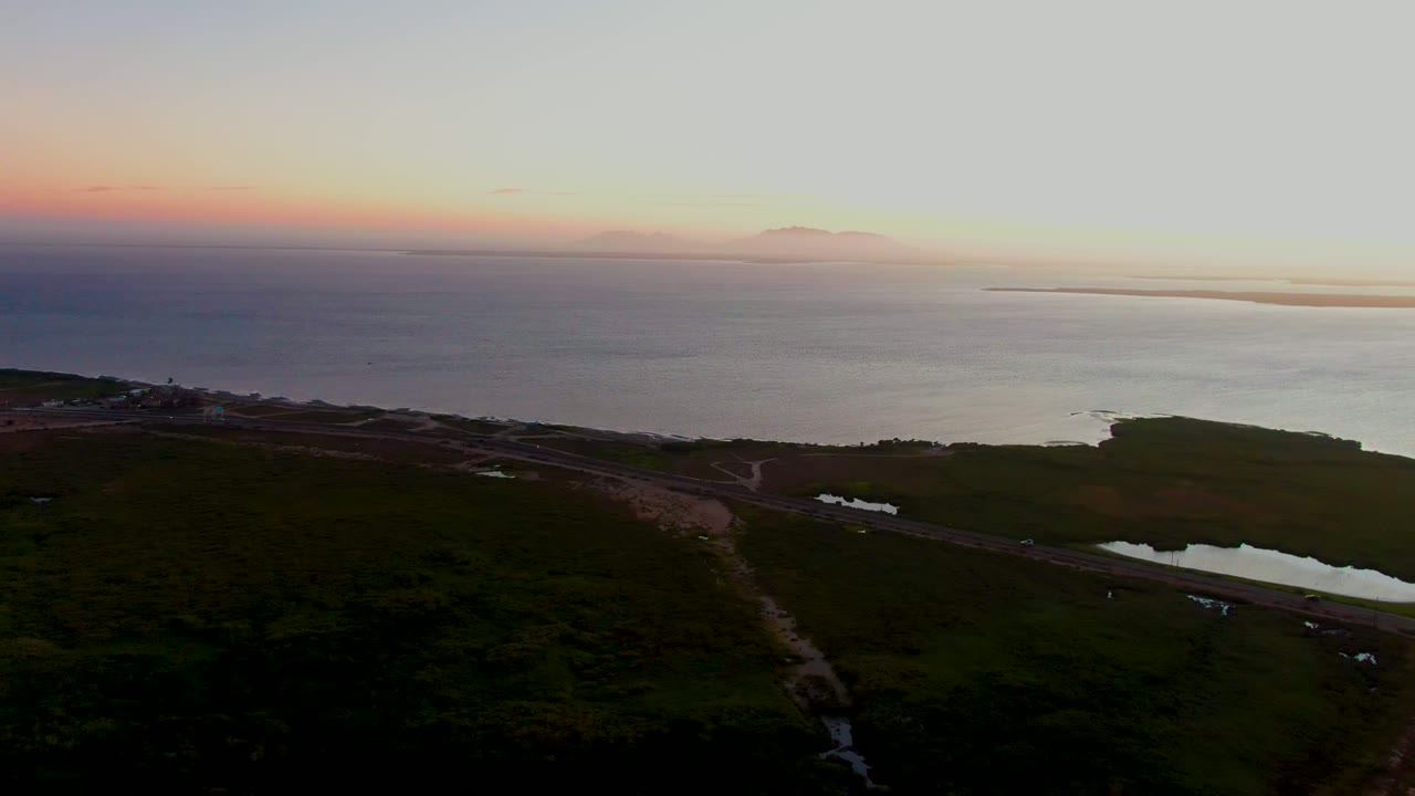 Aerial view in San Carlos, Baja California Sur with dark green fields below, a coastal road and pond in the foreground, ocean beyond, and no visible people.