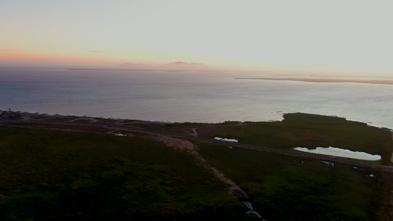 Aerial dusk view over San Carlos, Baja California Sur, with dark green coastal land, a road near small reflective ponds, open ocean, distant landforms, and no visible people.