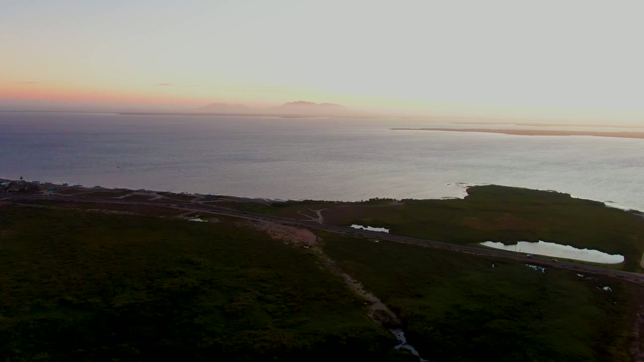 Wide aerial coastline with road, small ponds, dark fields, and warm horizon light.