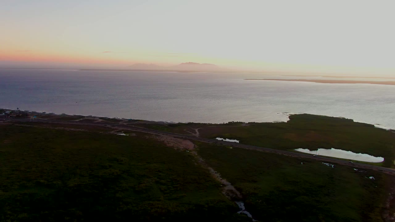 Aerial view over San Carlos, Baja California Sur, with a small vehicle on the coastal road, green foreground terrain, calm-looking ocean water, a warm horizon band, and no visible people.