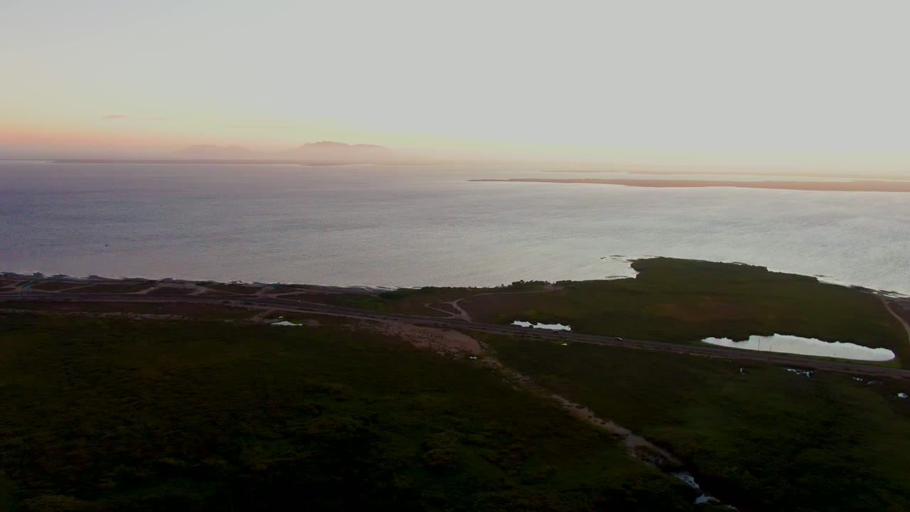 Aerial dusk view over San Carlos, Baja California Sur, showing dark grassland, a coastal path, reflective pools, open ocean, and no visible people.