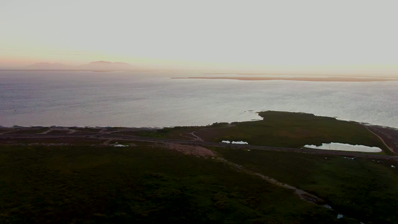 Aerial dusk view in San Carlos, Baja California Sur, showing green coastal land, a road near reflective ponds, calm water, a distant horizon, and no visible people.