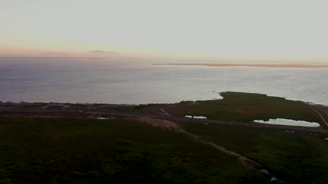 Wide aerial frame over San Carlos, Baja California Sur with muted pink and gray dusk sky, calm water, dark green land, a shoreline road, small ponds, and no visible people.