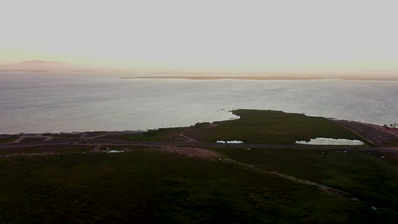 Aerial view over dark green coastal wetlands and small ponds near the calm sea in San Carlos, Baja California Sur, with a pale sunset horizon and no visible people.