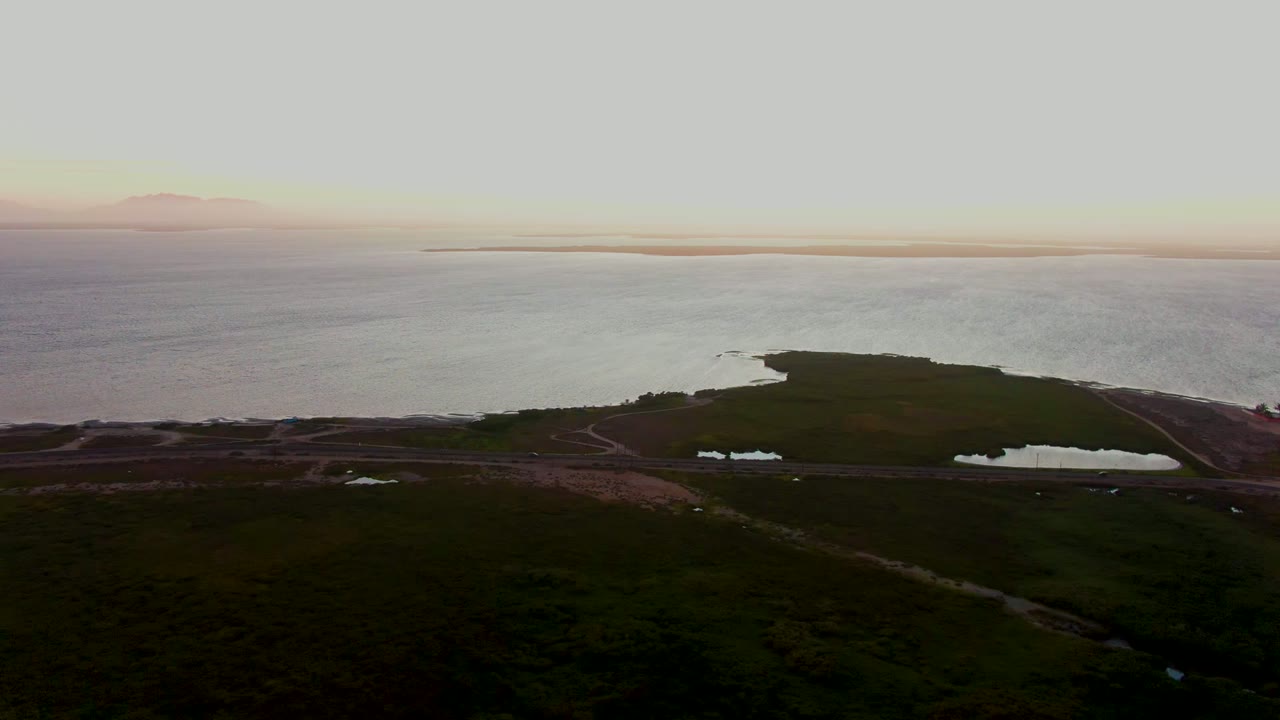 Aerial coastal landscape in San Carlos showing marshy green land in the foreground, small water bodies near a road, calm sea beyond, and no visible people.
