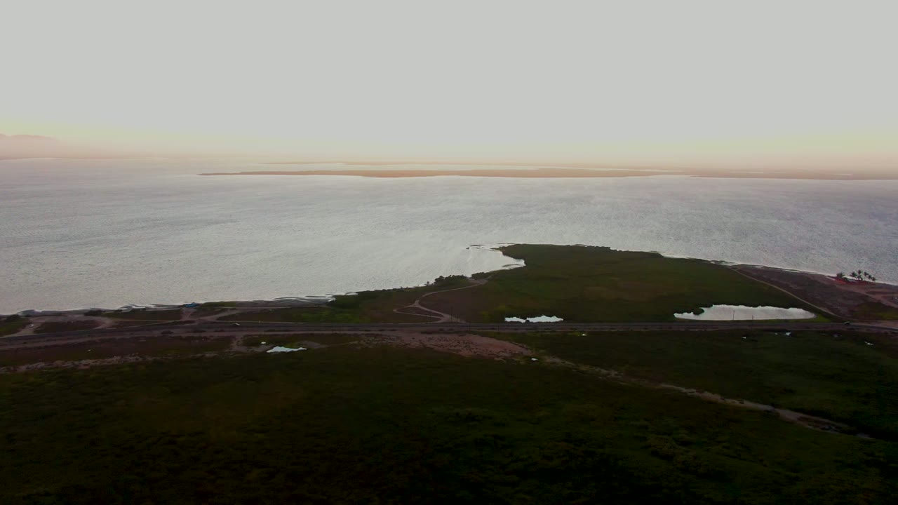 Aerial dusk view over San Carlos, Baja California Sur with dark green land in the foreground, a shoreline road and ponds near the coast, open water beyond, and no visible people.