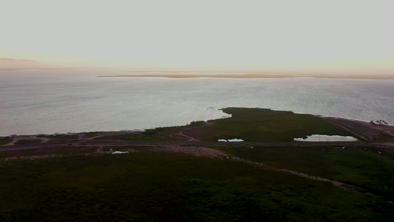 Wide aerial frame from San Carlos, Baja California Sur with a dark green peninsula shape, the shore road across the midground, small ponds, open ocean, and no visible people.
