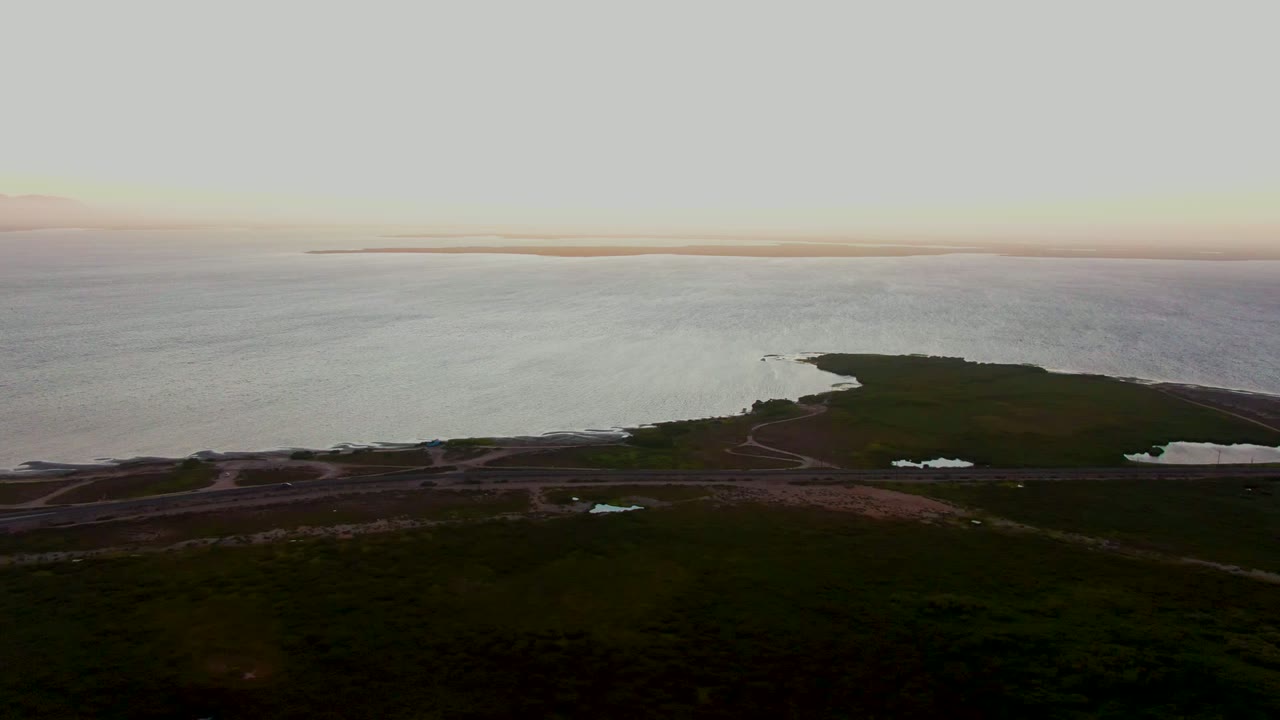 High aerial view over San Carlos, Baja California Sur, with dark green land in the foreground, calm coastal water beyond, a low road near the shore, and no visible people.