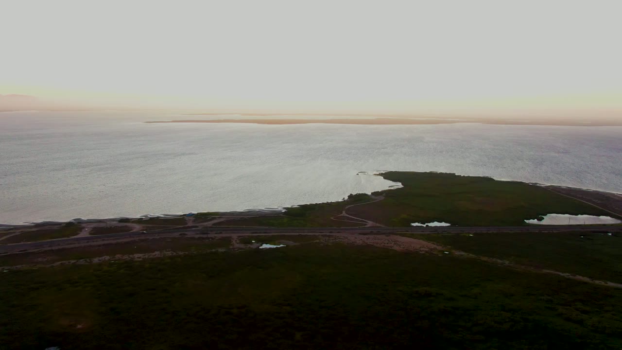 Wide drone view in San Carlos, Baja California Sur, with open coastal water behind a dark green strip of land, faint road curves near the shore, and no visible people.